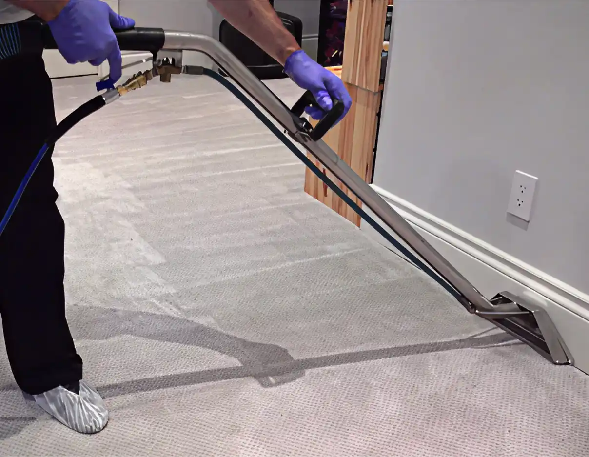 A carpet cleaning professional wearing blue gloves operates a steam cleaning wand to clean a grey carpet.